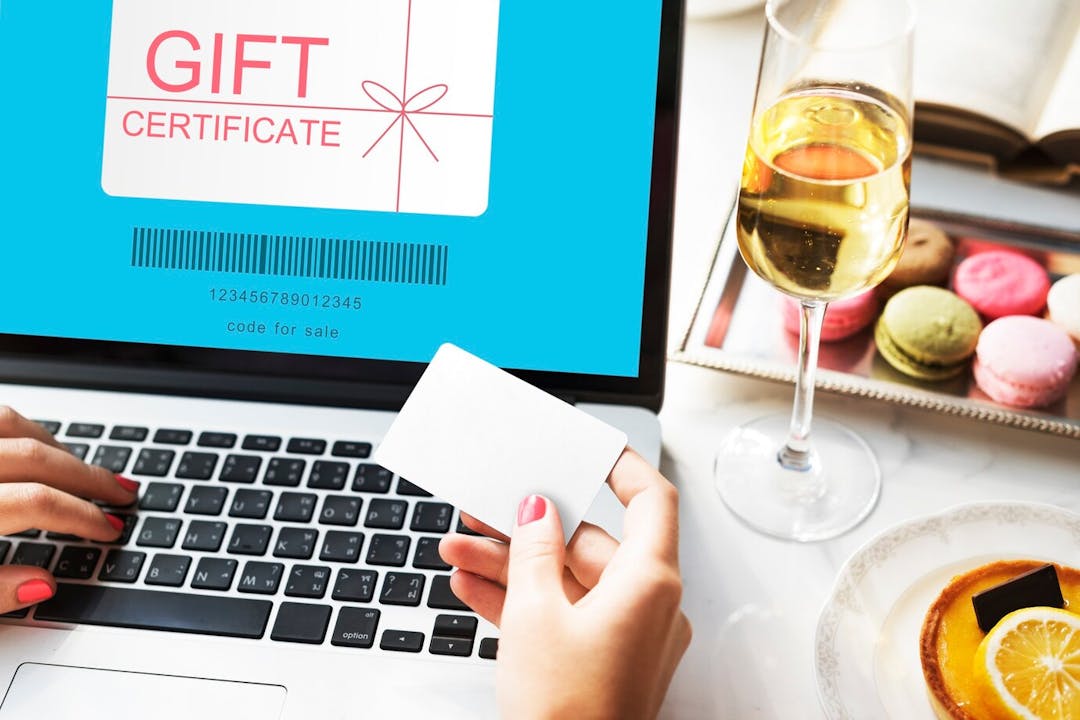 A person is holding a coupon near laptop and a glass of wine